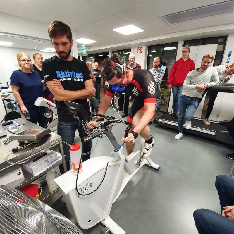 En person i atletisk utrustning och en mask cyklar på en stillastående cykel i ett labb medan människor observerar.