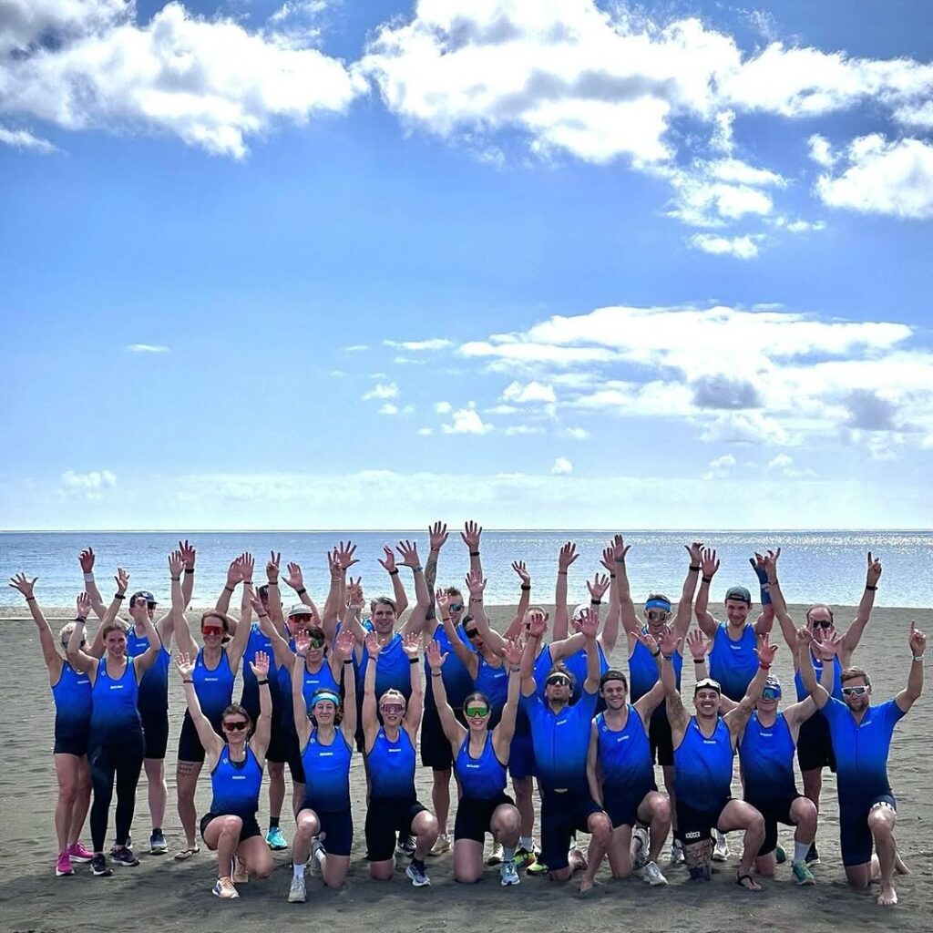 Grupp människor som bär blå toppar och svarta underdelar, poserar med upphöjda armar på en strand under en delvis molnig himmel.