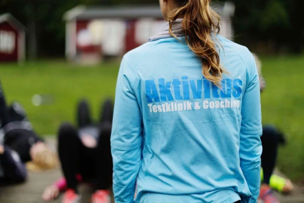 Person i blå sweatshirt med "Aktivitus Testklinik & Coaching" på ryggen står utomhus och observerar en grupp som tränar på marken. En röd och vit byggnad är suddig i bakgrunden.