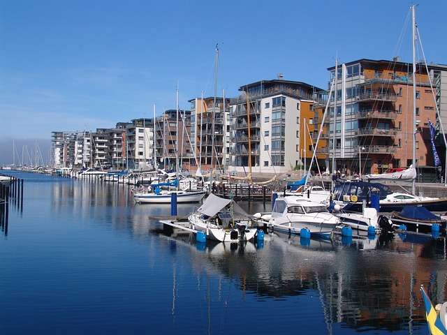 En rad segelbåtar lade till vid en marina med moderna hyreshus i bakgrunden under en klarblå himmel.