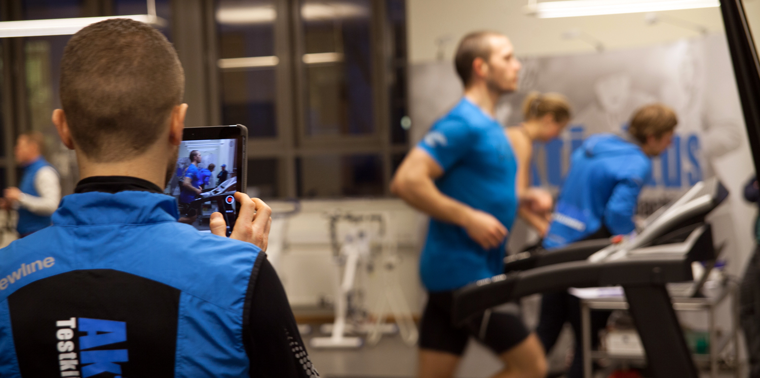 Människor som genomför och observerar ett konditionstest; en man springer på ett löpband medan en annan person spelar in processen med en surfplatta i ett gym.