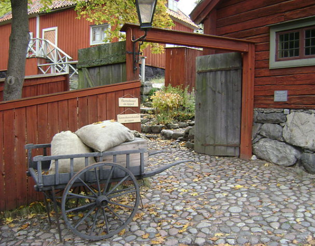 Trävagn med säckar på en kullerstensbana bredvid en röd träbyggnad och öppen port. Det finns en lampa och en skylt på staketet. Träd med höstlöv i bakgrunden.