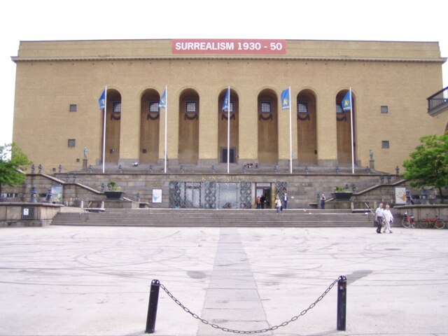 Stor byggnad med pelare och banderoller framför, med "Surrealism 1930-50". Trappsteg leder upp från ett brett torg, med några få personer som går.