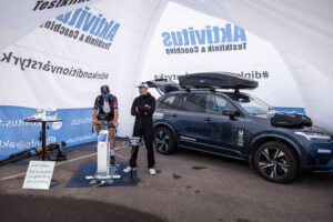 Cyklist på en stillastående cykel under ett märkestak bredvid en SUV. En annan person står bredvid dem. En skylt på ett stativ till vänster.