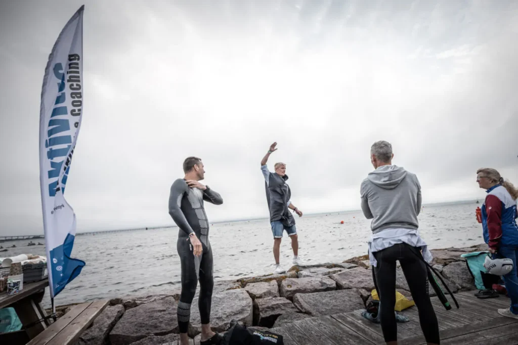 Fyra personer i atletisk utrustning står nära en stenig strandlinje under molnig himmel. En individ gester med en arm upplyft. En flagga med texten "OWMES Coaching" är synlig.