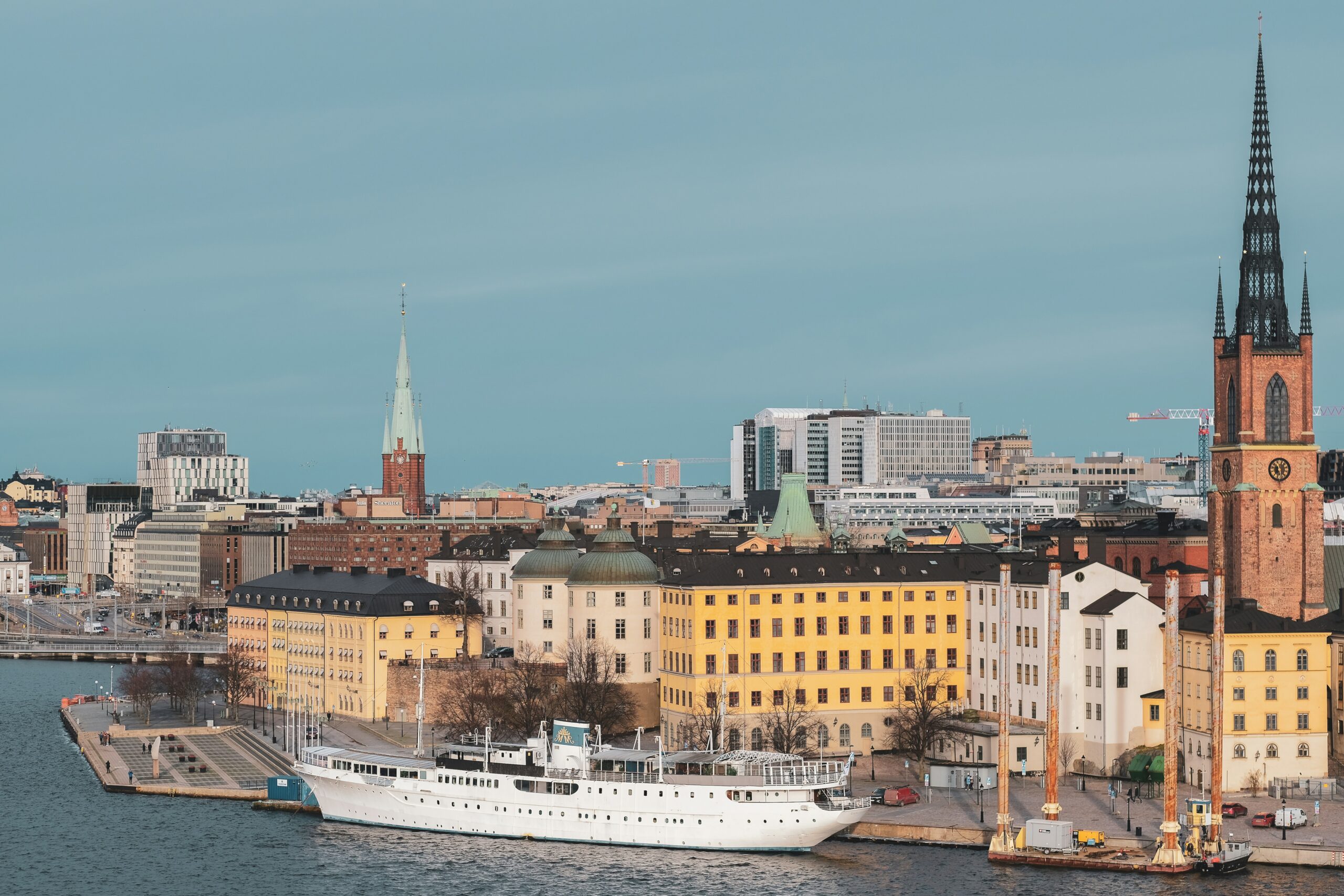 En kuststadsscen med ett dockat vitt skepp, historiska gula byggnader och höga spiror under en klar himmel.