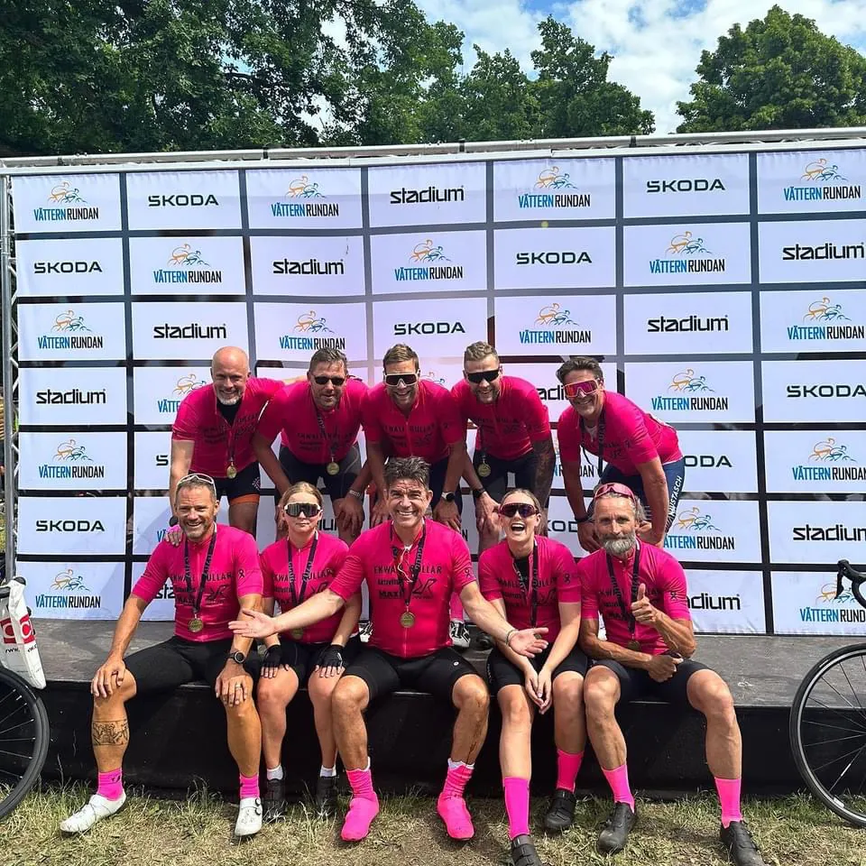 Grupp cyklister i rosa tröjor och medaljer poserar framför Vätternrundan i bakgrunden utomhus.