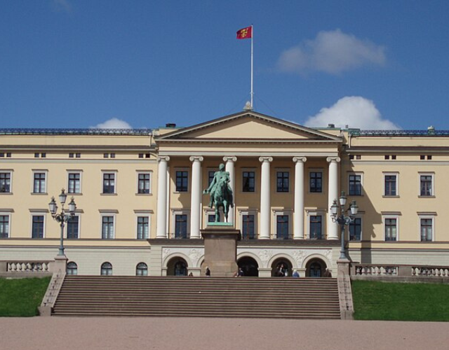 Stor beige byggnad med pelarfasad, en röd flagga på toppen och en ryttarstaty framför, mot en klarblå himmel.