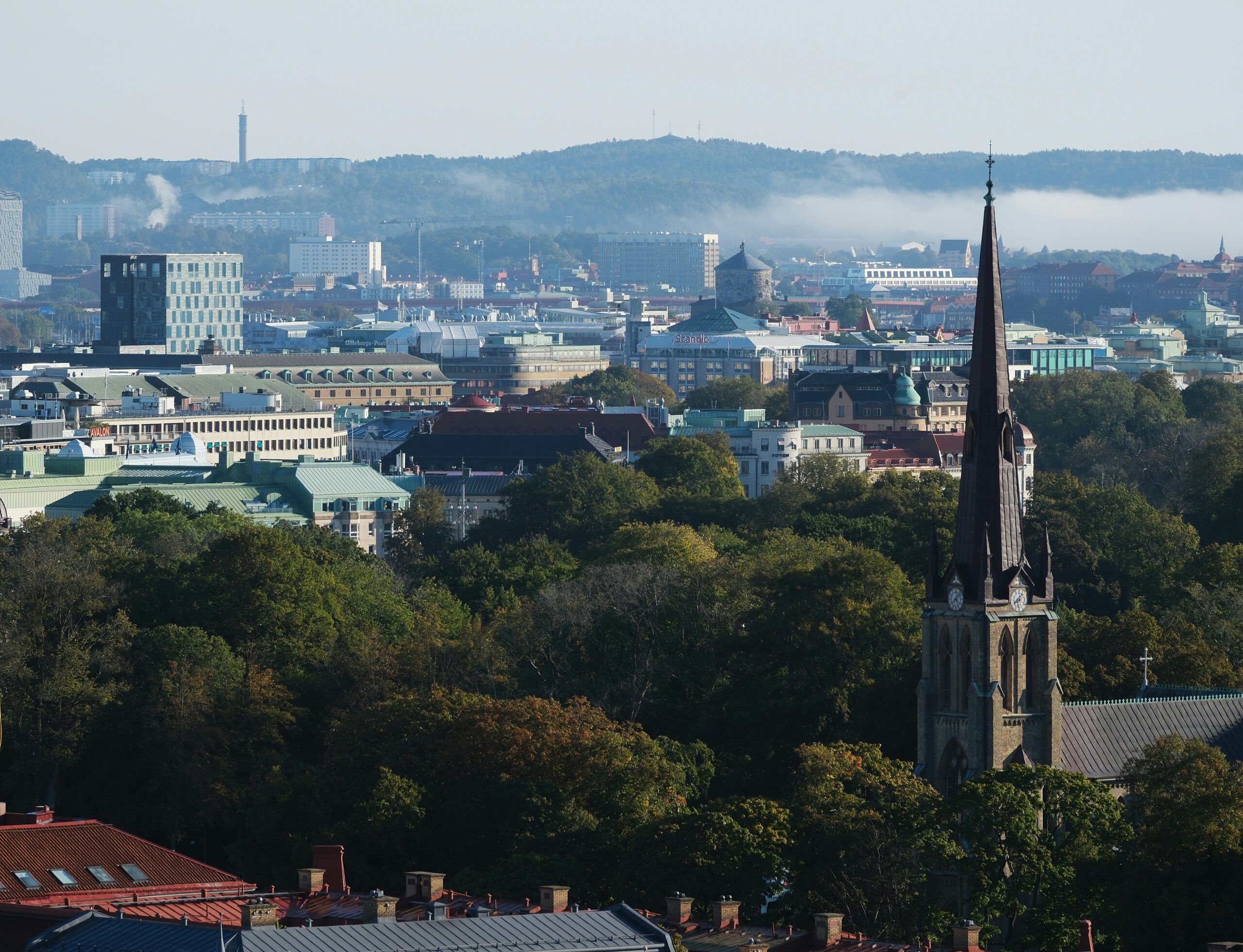 Flygfoto över en stad med en framträdande kyrkspira, omgiven av täta gröna träd, med en bakgrund av avlägsna kullar och spridda byggnader.