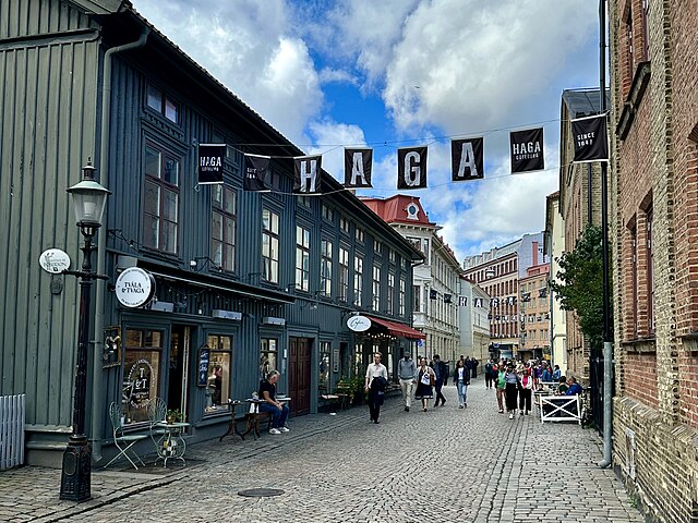 Kullerstensgata i Haga, Göteborg, kantad av träbyggnader, butiker och caféer. Människor går längs gatan under banderoller. Molnig himmel ovanför.