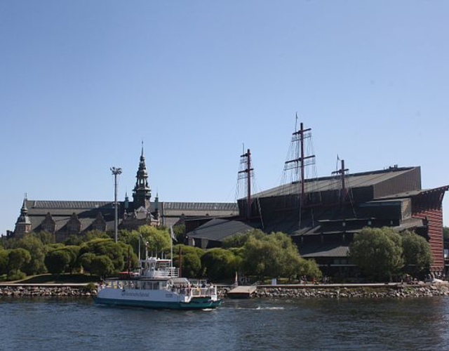 Färja på en flod med en utsmyckad byggnad och ett skeppsmuseum i bakgrunden, under en klarblå himmel.