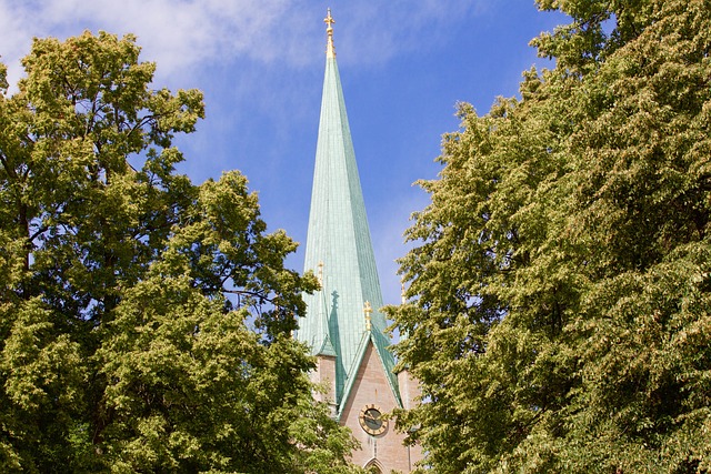 Ett grönt kyrktorn med ett guldkors reser sig mellan höga träd under en klarblå himmel.