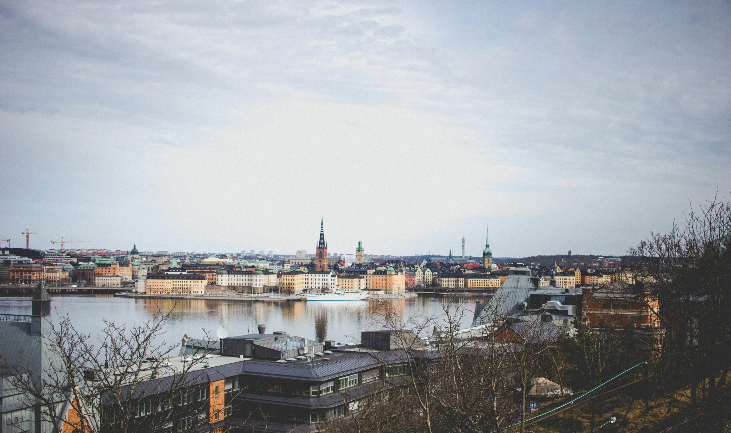 En stadsbild har en strandpromenad med historiska byggnader och flera kyrkspiror under en molnig himmel. Kala träd syns i förgrunden.