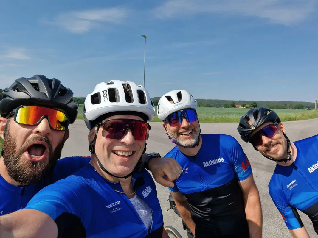 Fyra cyklister som bär blå tröjor och hjälmar poserar för en gruppselfie en solig dag. Ett lantligt landskap med en klarblå himmel är i bakgrunden.