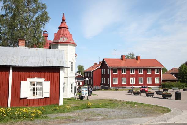 En byscen med röda trähus, inklusive ett med ett högt torn, en central innergård, bänkar och gröna träd under en klarblå himmel.