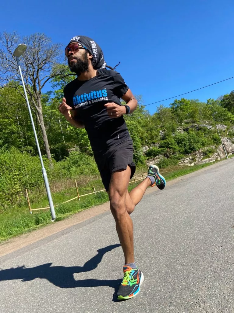 En man klädd i svart skjorta och shorts joggar på en asfalterad väg i ett skogsområde under klar himmel.